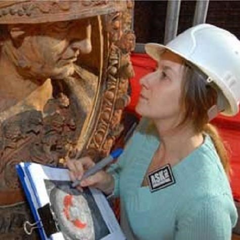 A woman in hard hat, with a clip board, stares at a statue.