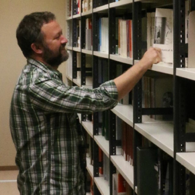 A man in plaid shirt takes a book off a shelf