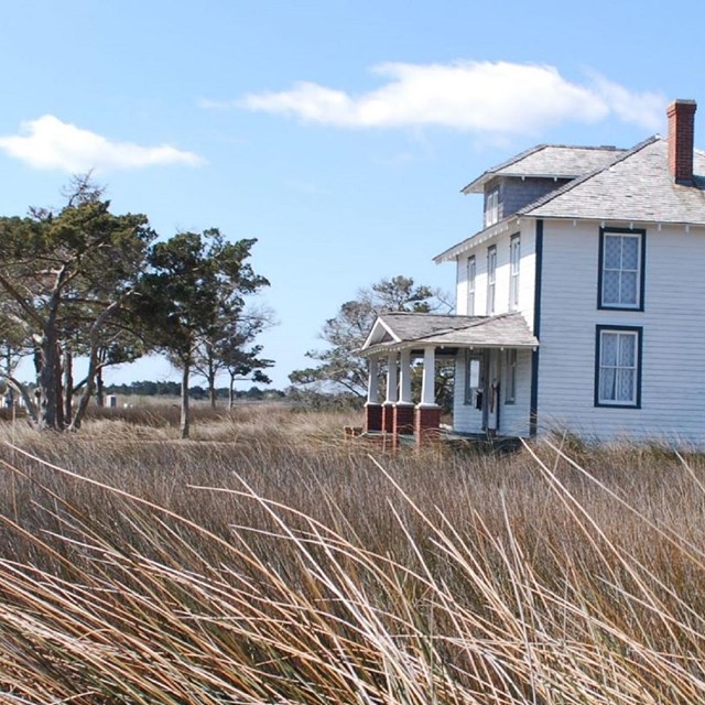 A white clapboard house sits on the right with brown grass and two trees in front.