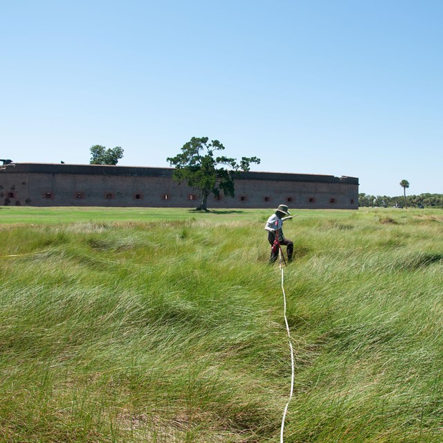 Woman holding a measuring tape waking in tall grass in front of fort.