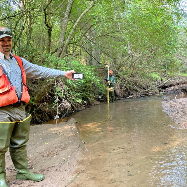 Man in orange vest and waders holds a card with a number, Man holding survey equipment in background