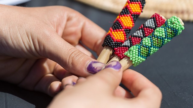 A close-up of two hands holding three short sticks wrapped in colorfully-patterned beads