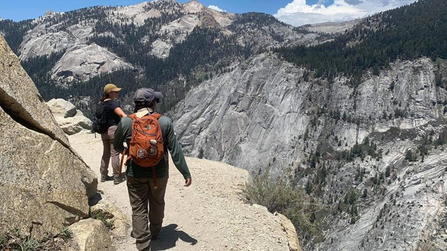 Hikers walk on a gravelly trail next to a steep cliff with rugged mountains in the background. 