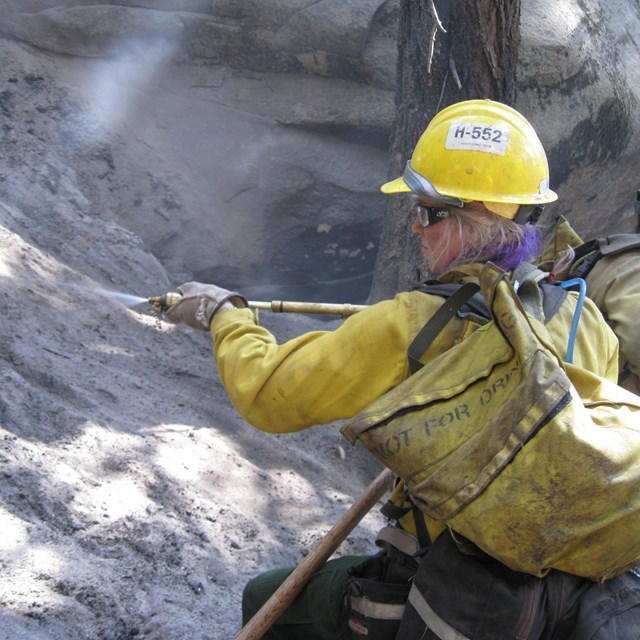 A woman in a yellow shirt and hardhat sprays water into a hot, ashy landscape.