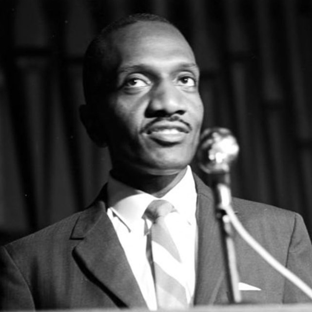 Black and white photo of a black middle-aged man speaking at a podium.