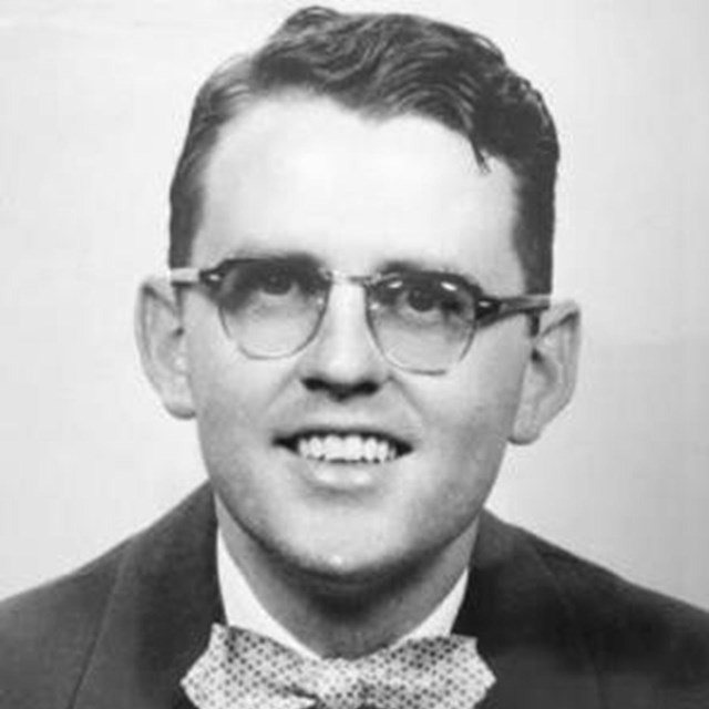 Black and white portrait of a smiling white man with glasses wearing a suit and bowtie.