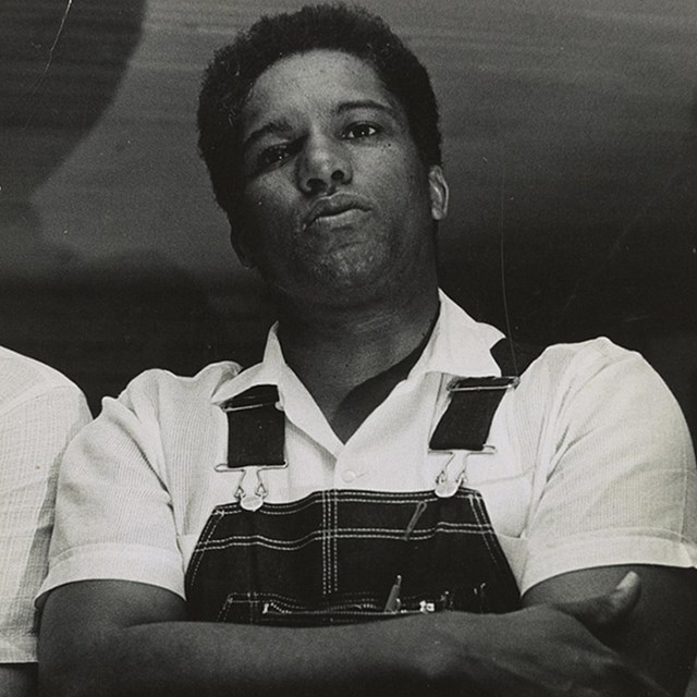 Black and white photo of a young black man wearing overalls and standing with his arms crossed.
