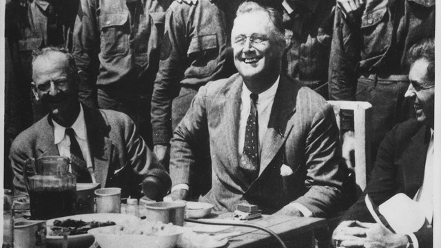 A black and white image of President Hoover sitting at a dinner table with a group of CCC boys.