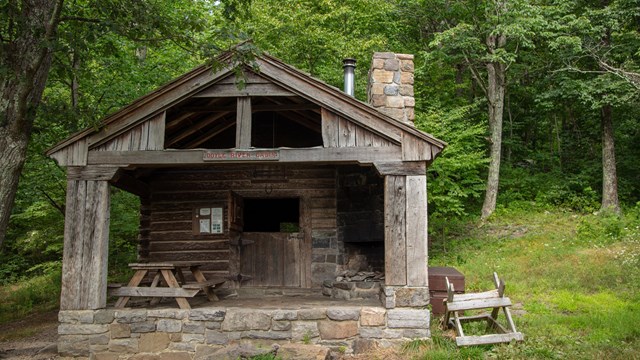 Log cabin with stone fireplace and chimney. A picnic table sits on the stone porch.