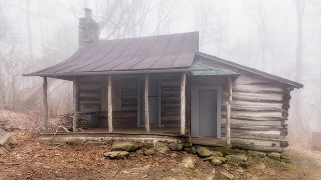 A wooden rustic cabin with a porch, two doors, and a stone chimney is surround by fog in the woods.