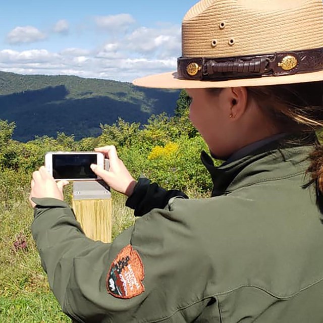 A park ranger takes a picture of a mountain overlook with their cell phone.