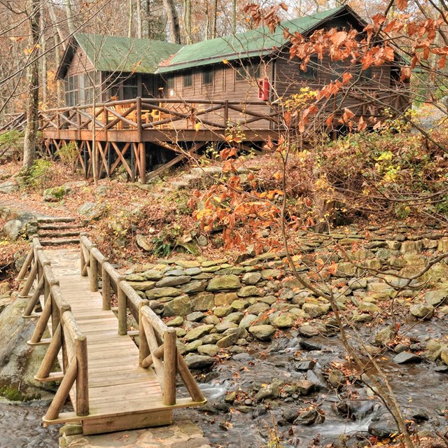 A historical color photograph of a log cabin with a bridge crossing over a creek in the foreground.