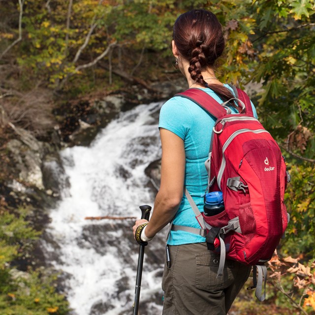 A woman hiker with a red backpack looks out over a waterfall in the distance.