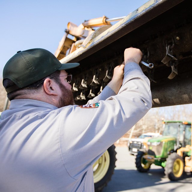 A man in a park ranger uniform works on a tractor.