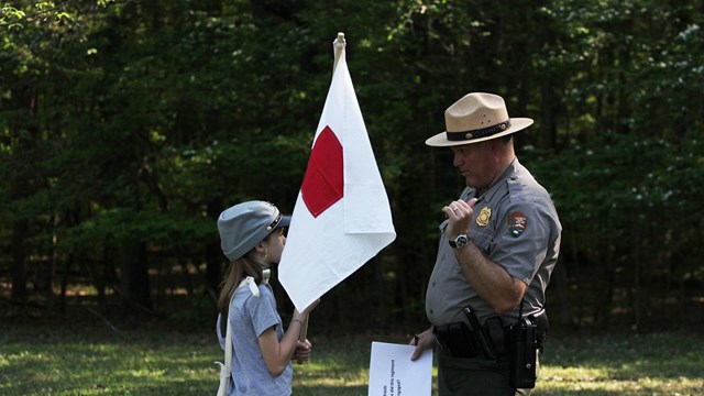 A young girl holding a flag listens to instructions from a Park Ranger. 