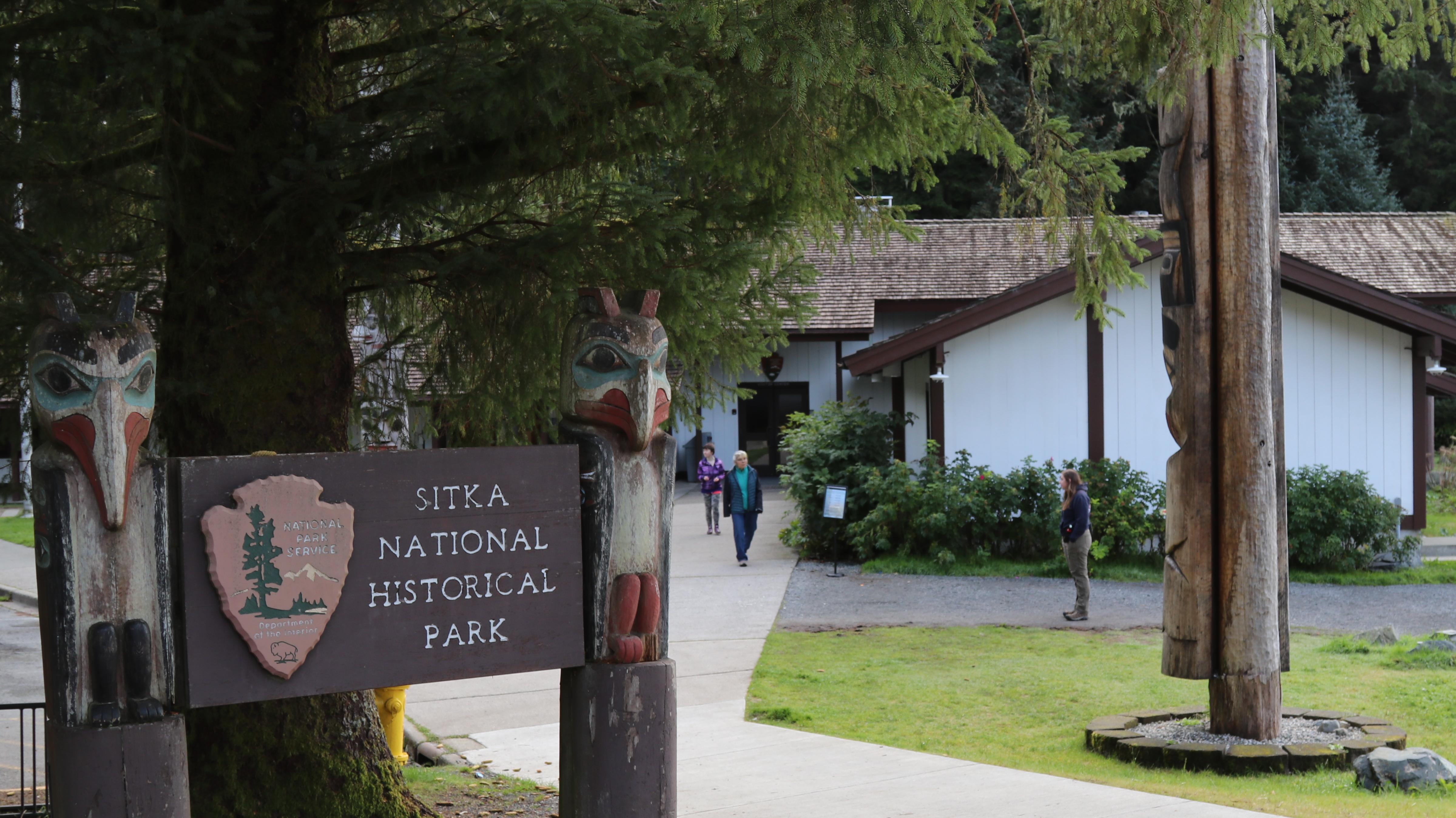 Park entrance sign in front of a tree. people on sidewalk leading to a totem pole and visitor center