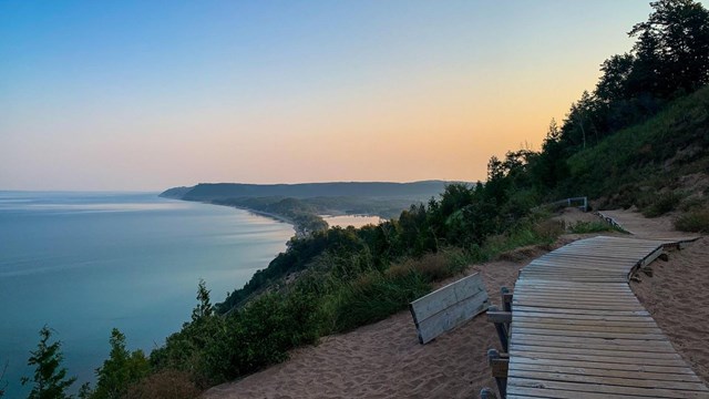 Looking down the bluff, a sunrise lights the sky in rainbow of colors and a bright blue lakeshore.