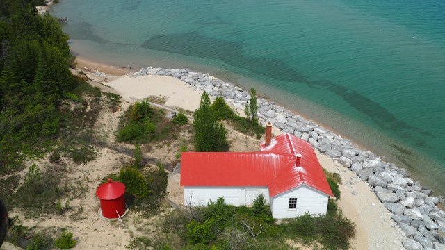 Looking down, bright red roofed building at the edge of a rocky shore bleeds into a blue lake. 