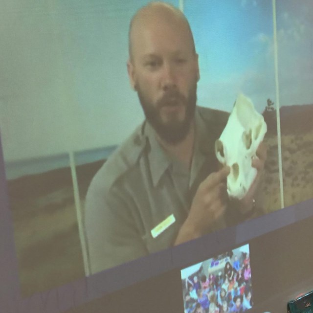Ranger on a tv screen holds up a skull during a distance learning program