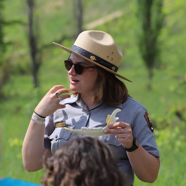 A park ranger wearing a flat hat gestures toward her mouth while holding a deer jaw