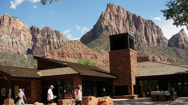 People walk park a visitor center with towering rock formations in the background.