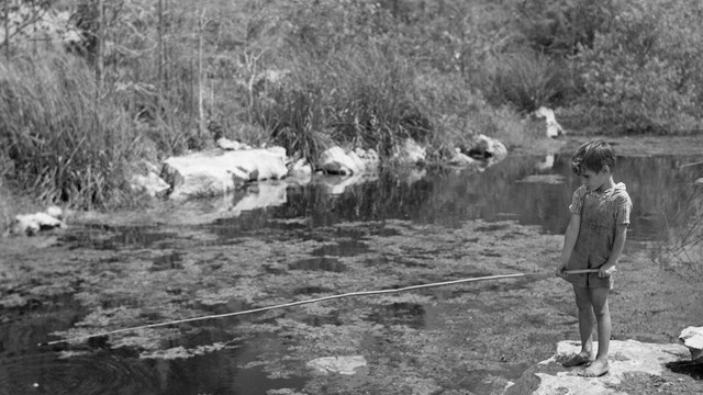 A young boy barefoot standing on the edge of water on a rock while fishing.