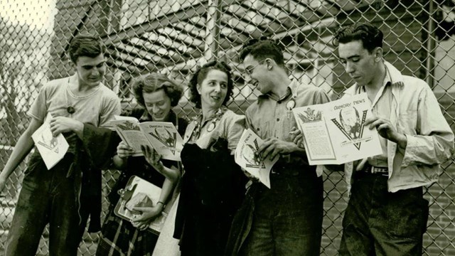 A black and white photo of 5 people holding magazines standing along a fence.