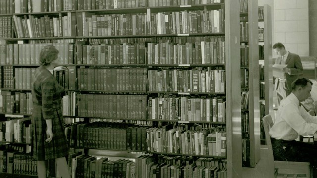 A black and white photo of a woman standing in front of a library shelf full of books. 