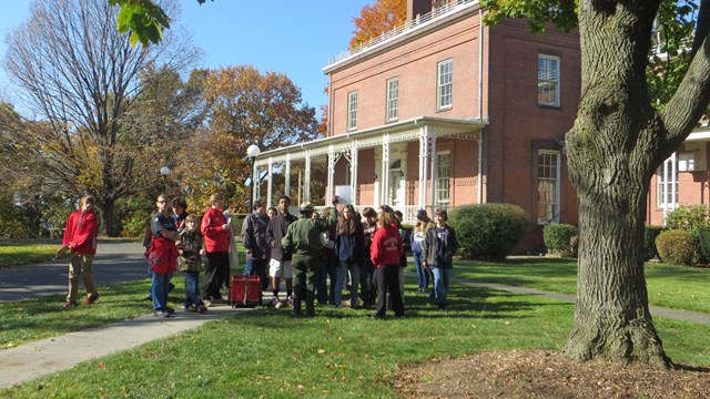 A group of students stand with a park ranger outside of a building with a white porch. 