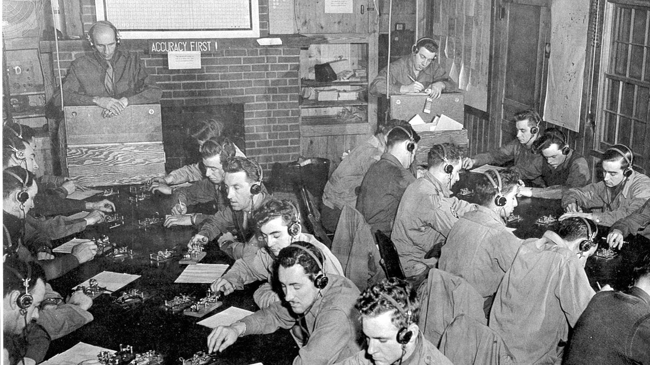 B&W photo of men seated in rows at tables in rustic room with headphones on