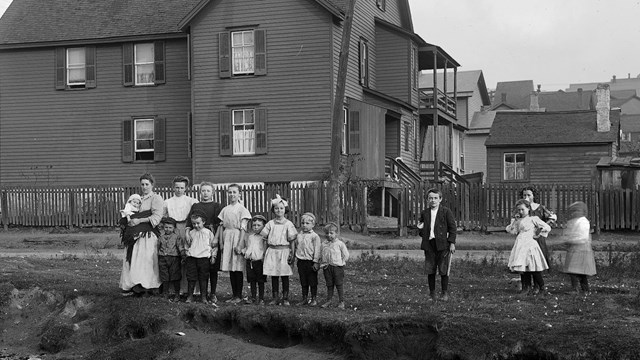 A black and white photo of a family of 15 women and children posing in front of a house by a runoff