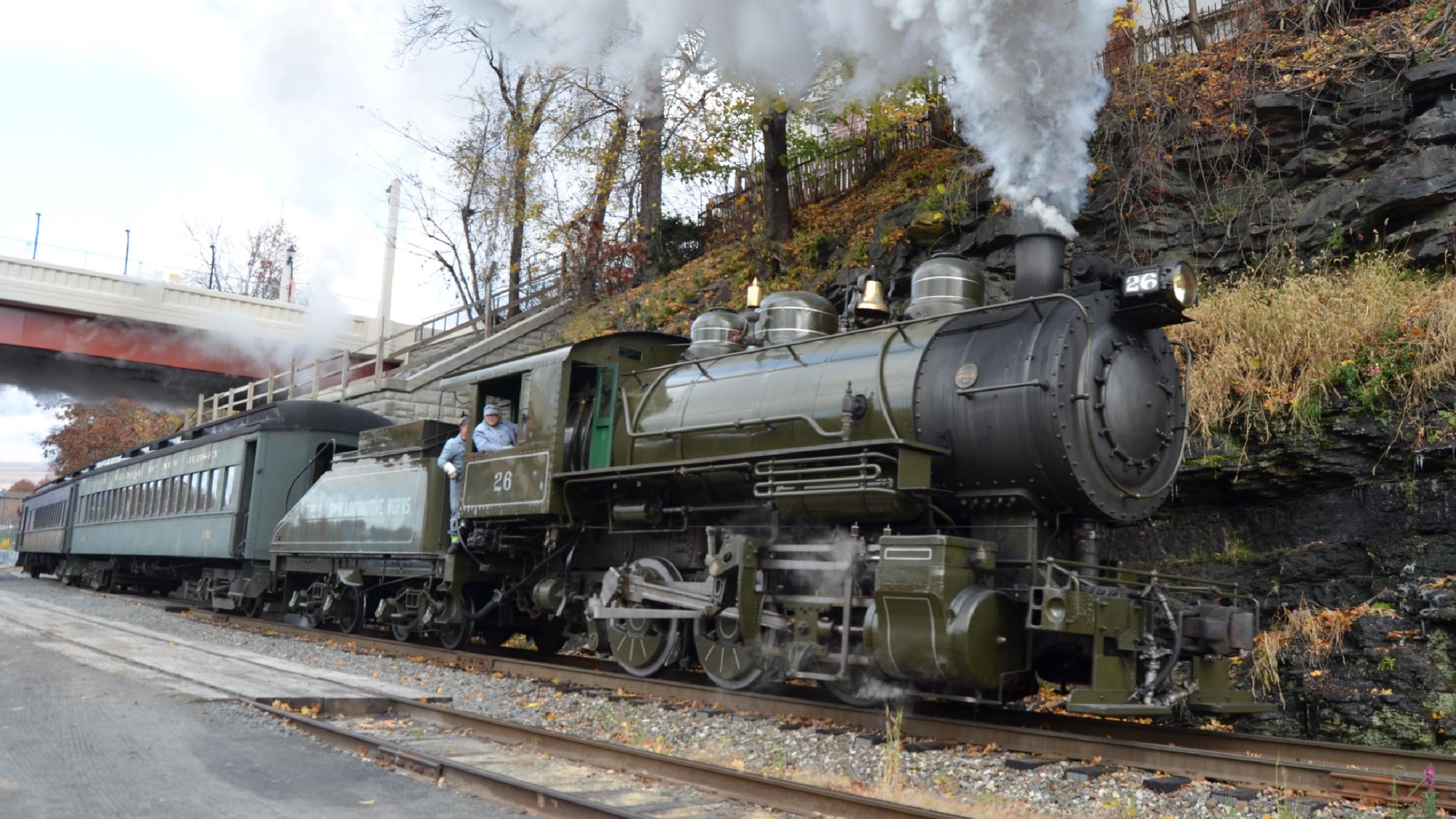Steam locomotive running on a track under a bridge with a woodsy hillside in the background