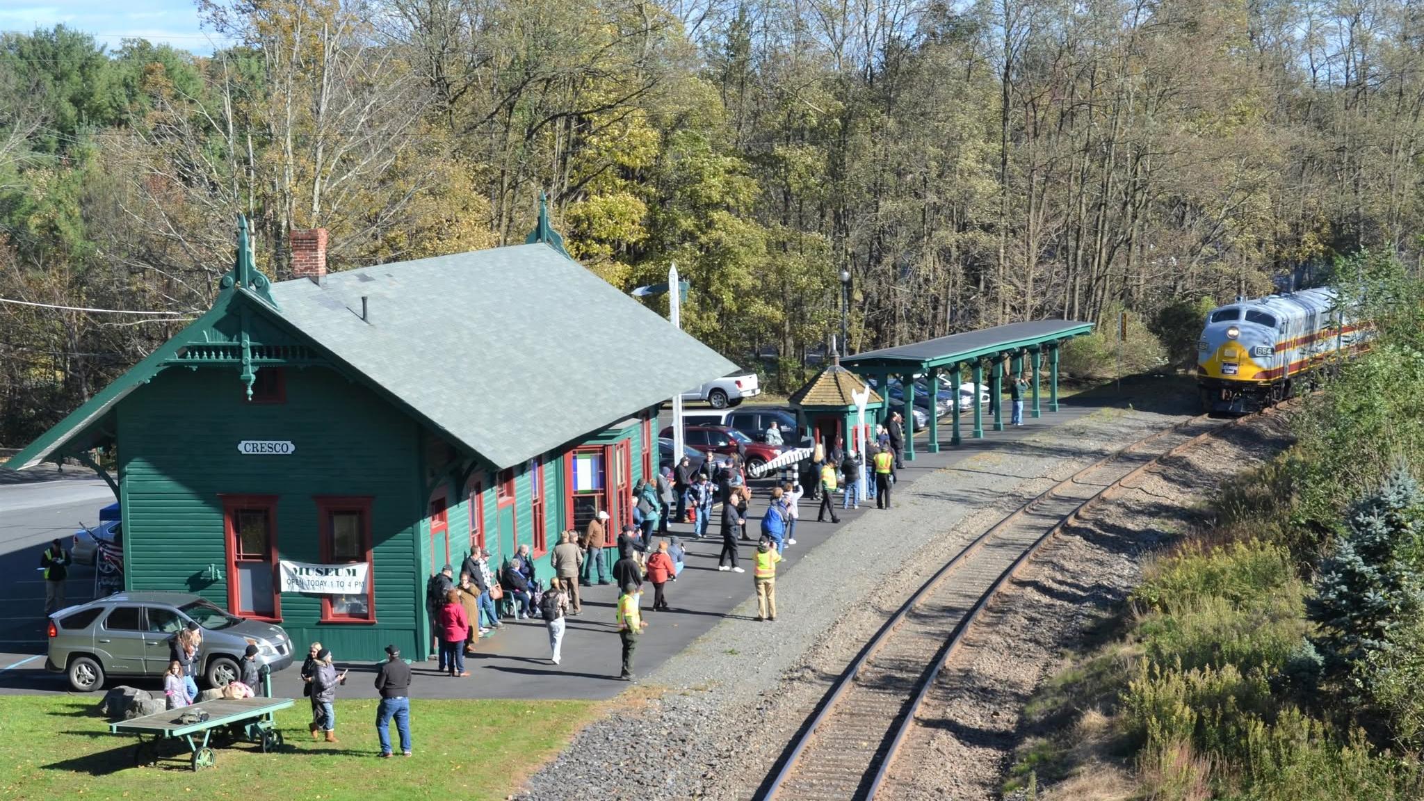 A classic 1940s locomotive approaches a crowded green railroad depot