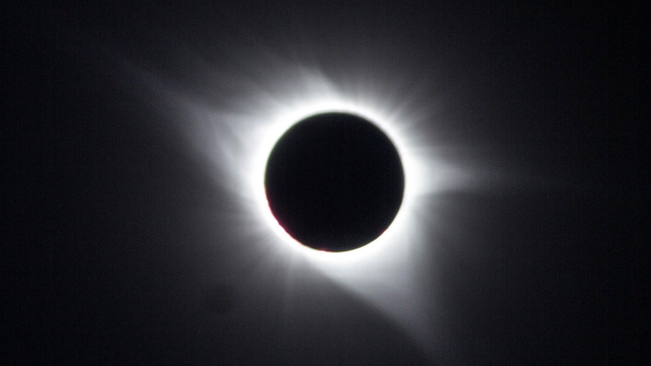 White corona of the sun is visible around black circle of the moon in a dark sky.