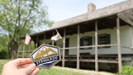 A hand holding an embroidered patch that reads Stewards. A historic home is in the background.