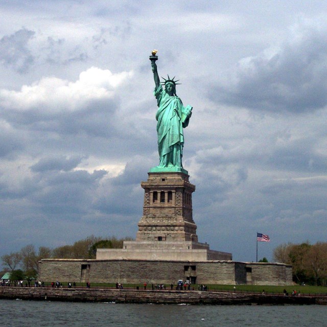 The Statue of Liberty from the water. The green color of the statue is vivid against a cloudy sky.