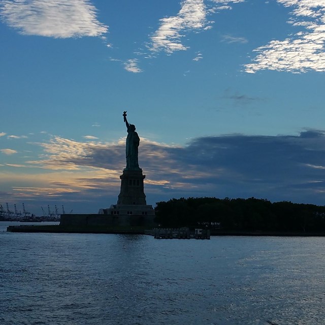 The Statue of Liberty and Liberty Island against a sunset in the harbor.