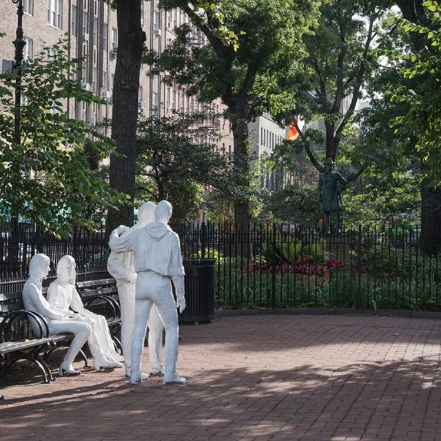 Christopher Park, surrounded by green benches, laid with brick, with two white statues in the center