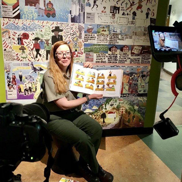 A park ranger sits in front of a camera recording her as she reads from a picture book