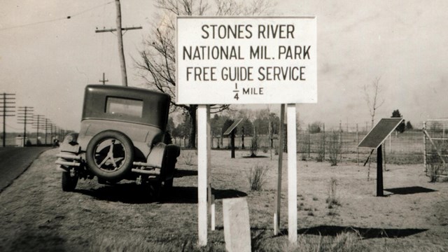 Black and white image of Stones River road sign with early model car from 1930s.
