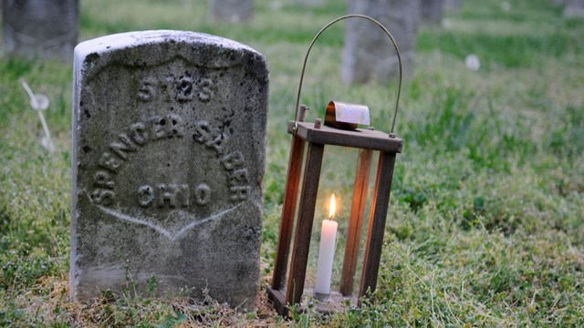 Gravestone with text reading S123 Spencer Sable Ohio and lantern with lit candle to the right
