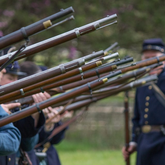 A row of soldiers hold muskets in the air with a Union soldier looking on in the background.