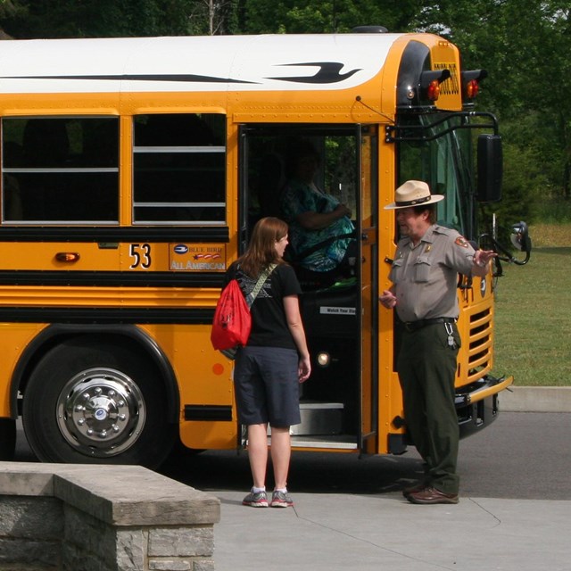 A ranger in uniform speaks to a teacher in front of a school bus.