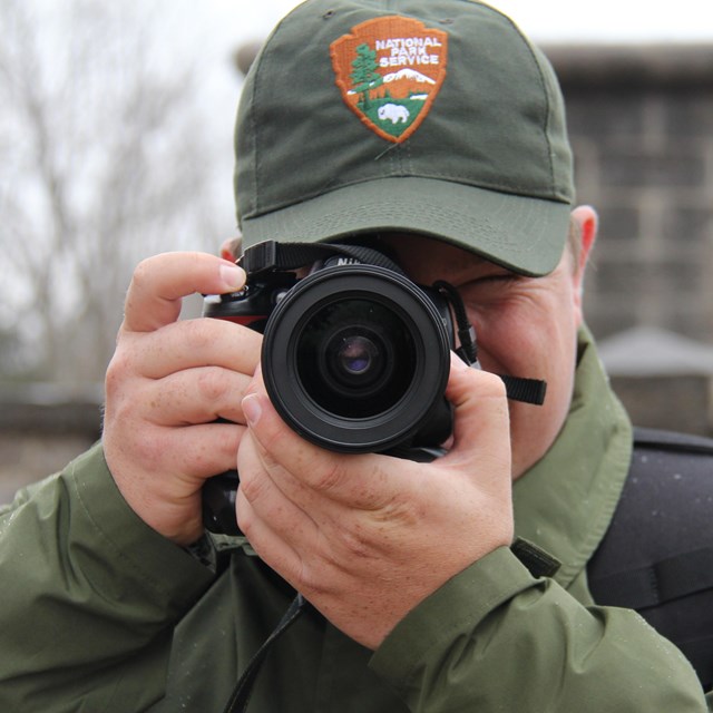 A Park Ranger in uniform holds a camera up to his eye and points it at the viewer.
