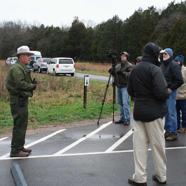A park ranger in uniform speaks to a group of people, one of which with a video camera
