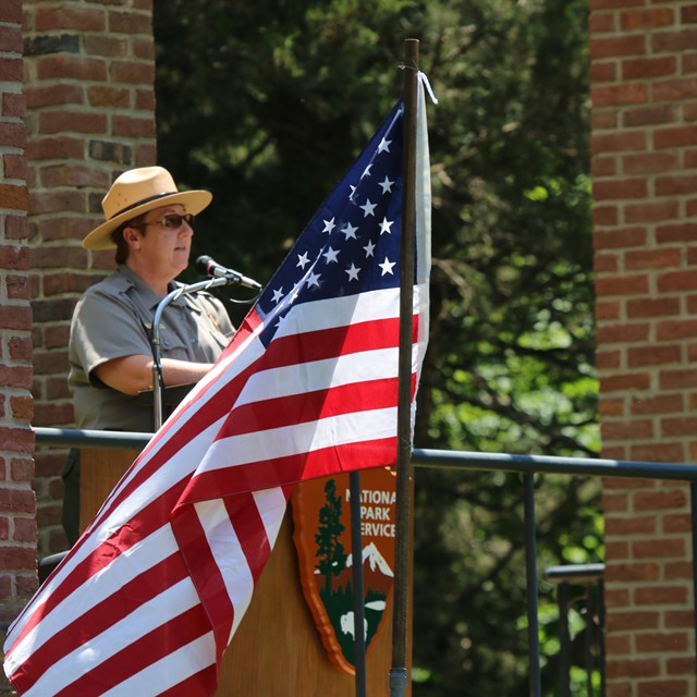 A park ranger in uniform speaks at a podium with an American Flag in front of her.