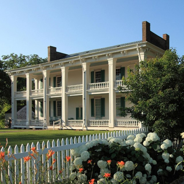 A white house with seven columns and two chimneys. In the foreground is a white fence and flowers.
