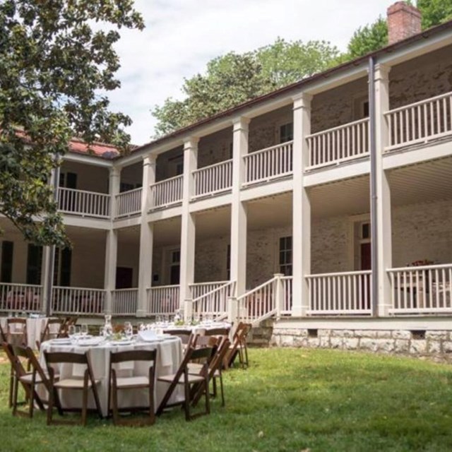 Several tables are set out in front of a old two story brick home with a balcony and many columns.