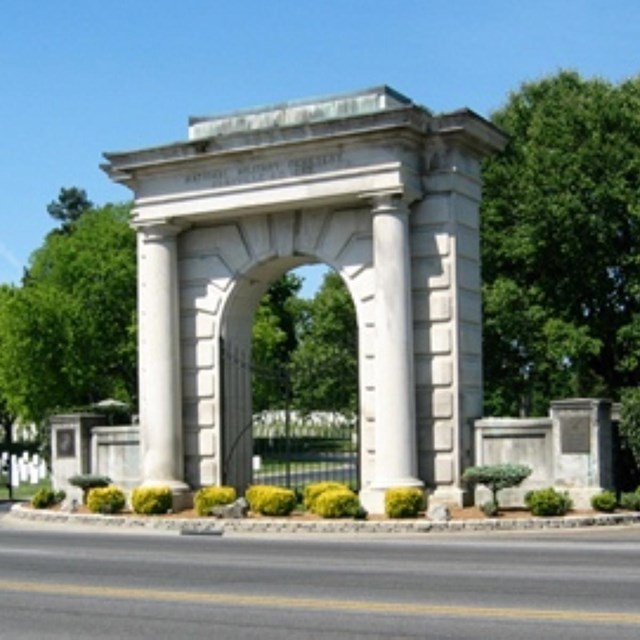 A stone arch with a iron gate into a cemetery with entrance and exit roads on each site.