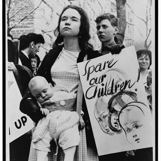 Woman wearing baby and holding sign reading Spare Our Children